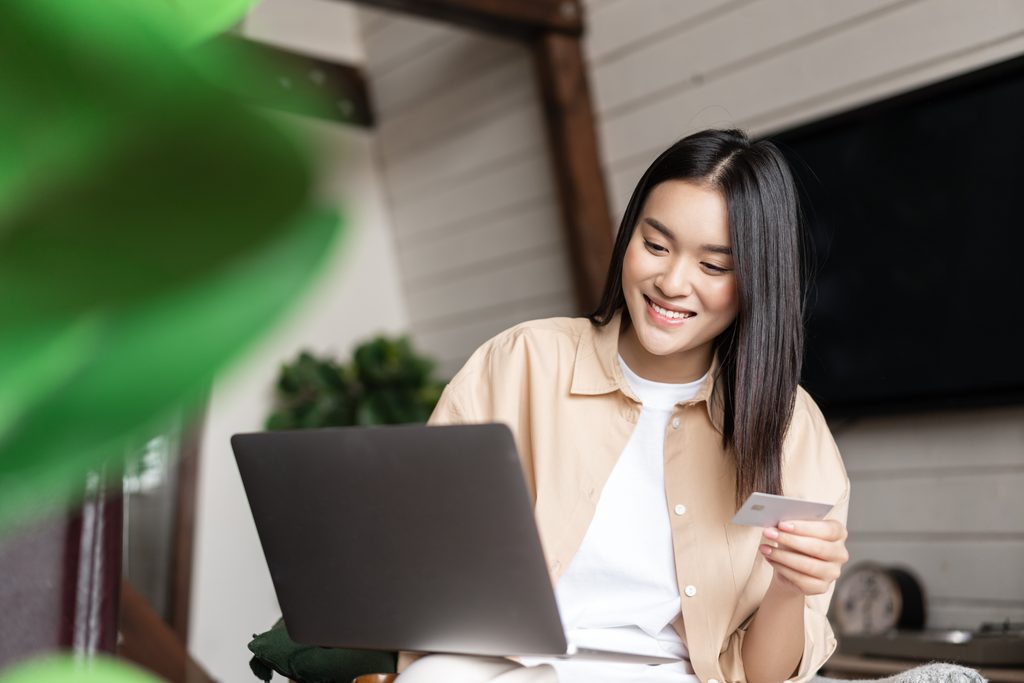 Image of korean woman buying smth online, paying with credit card on website, purchase on laptop from home.