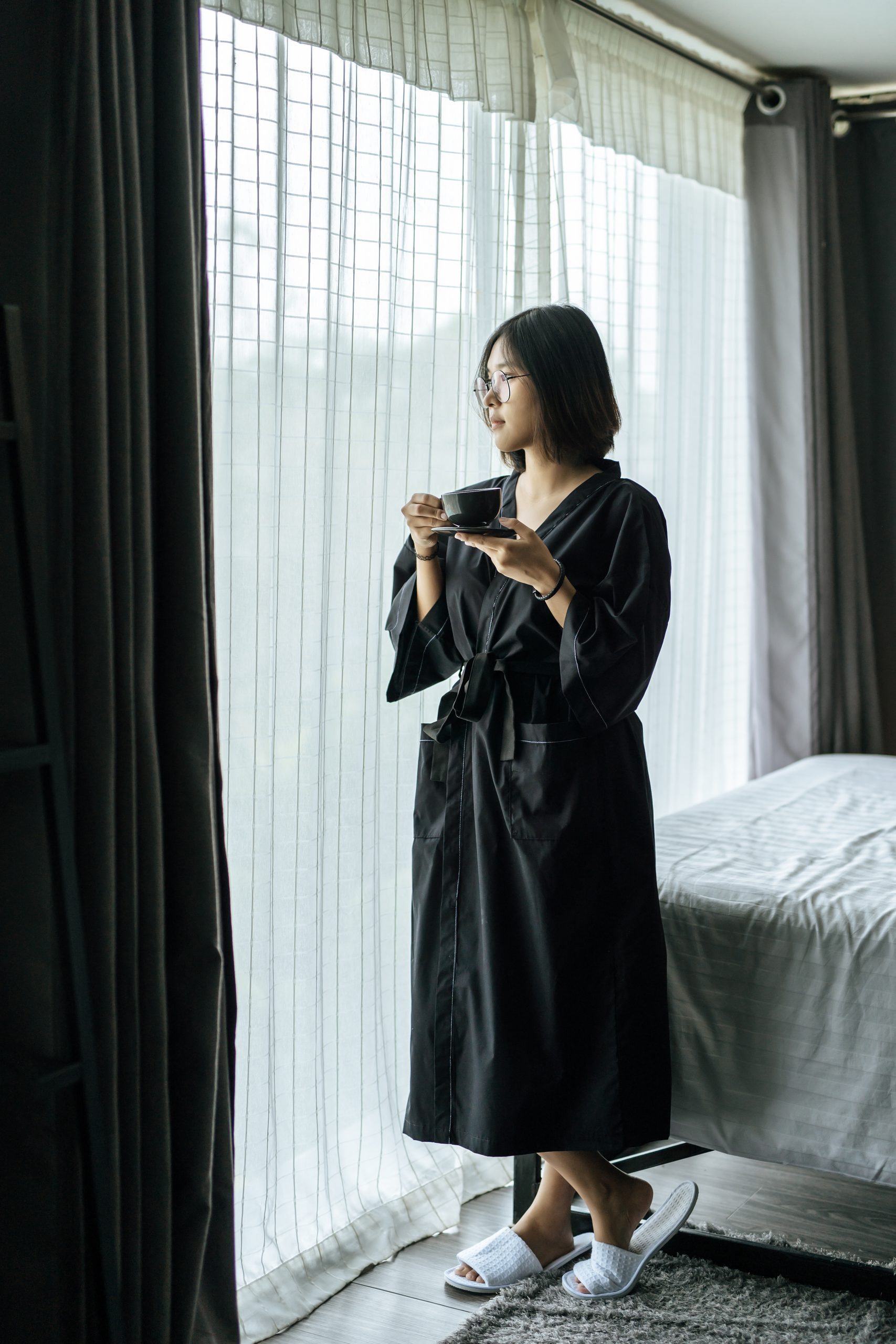The woman wore a black robe near the window and drank coffee in the bedroom.